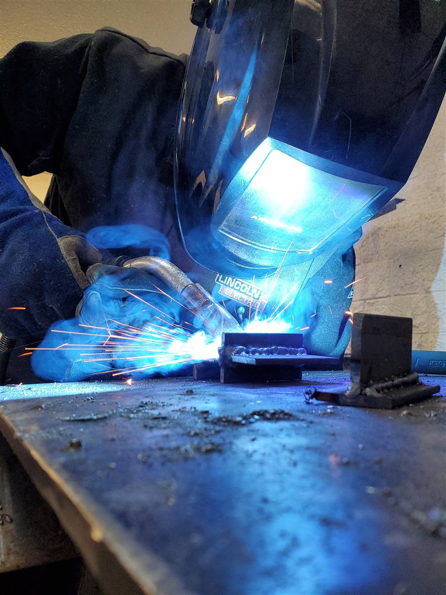 Welder MIG welding a fillet joint with flying sparks, with a finished T-joint beside the plate