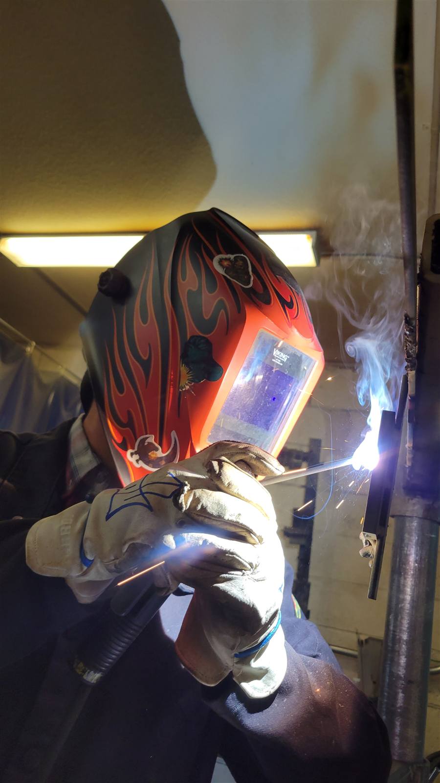 Welder in a flame-decorated helmet stick welding a vertical joint, with smoke and sparks